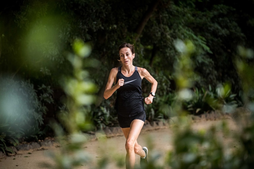 A woman in her 40s runs in front of greenery on a gravel path.