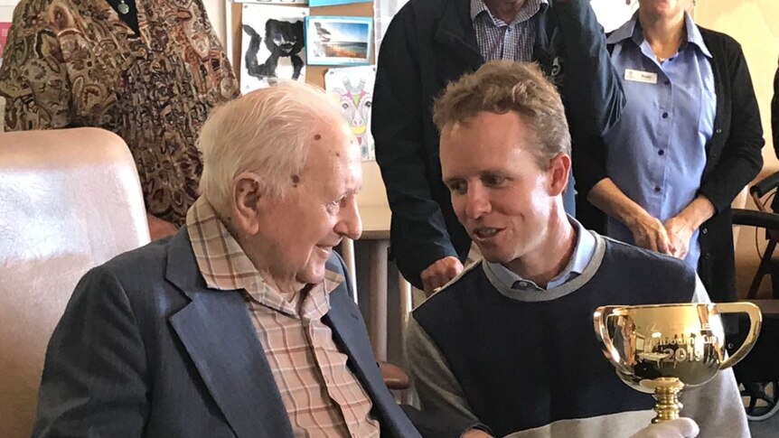 An elderly man sits listening to a younger man talk as he holds the golden Melbourne Cup