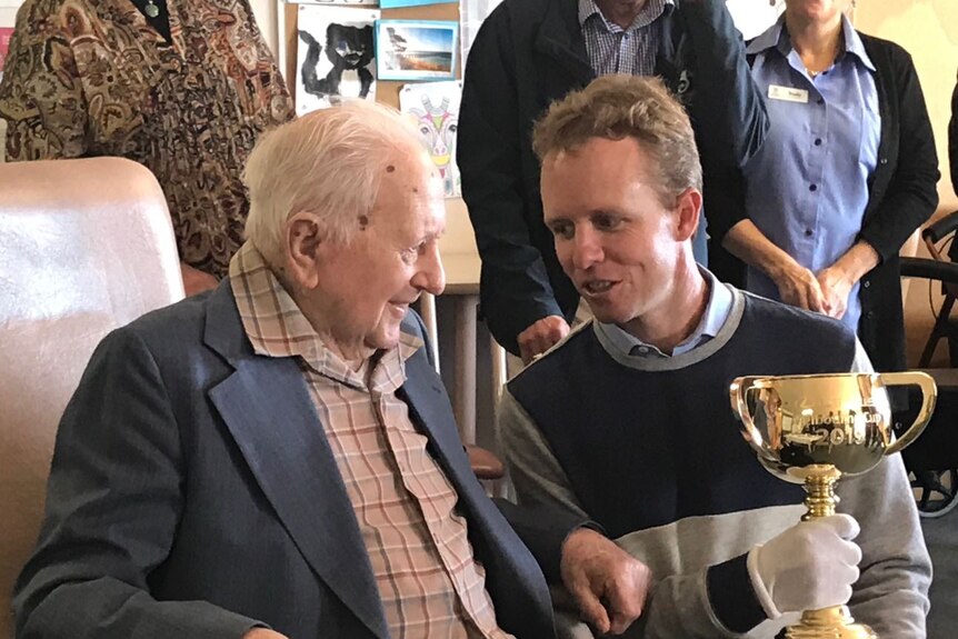 An elderly man sits listening to a younger man talk as he holds the golden Melbourne Cup