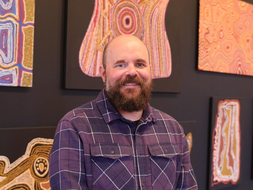 Glenn smiling while pictured in front of Indigenous artwork wearing a flannelette shirt