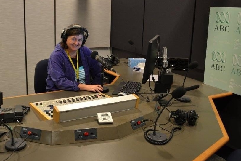 Woman wearing headphones sitting at desk in radio studio in front of microphone and audio desk.