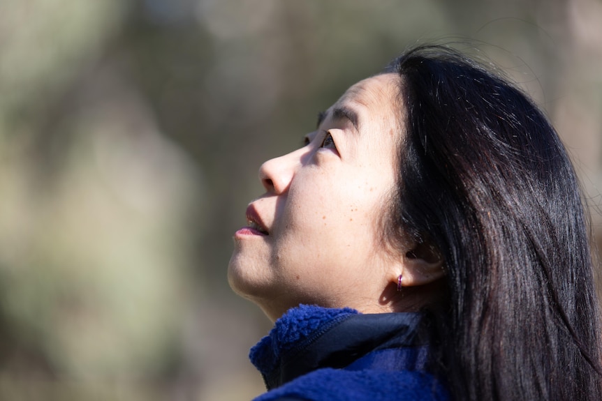 Woman with black hair with a happy look on her face looks up into trees.