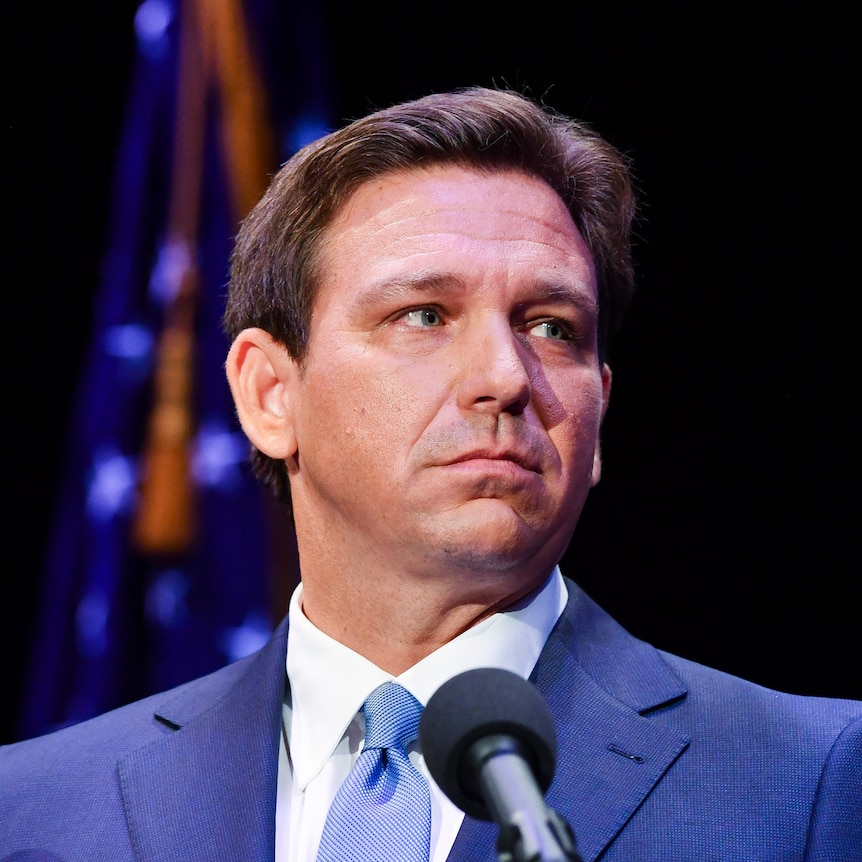 Ron Desantis looks to one side while standing behind a microphone and wearing a blue suit