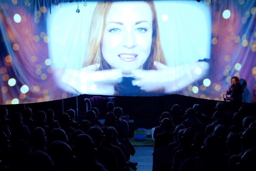 A projected image of a woman face on a stage with people's heads visible in the audience.