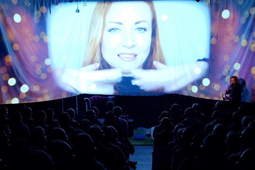 A projected image of a woman face on a stage with people's heads visible in the audience.