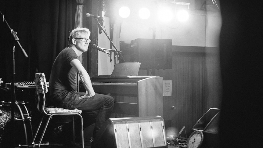 Black and white photo of David Bridie sitting at a piano 