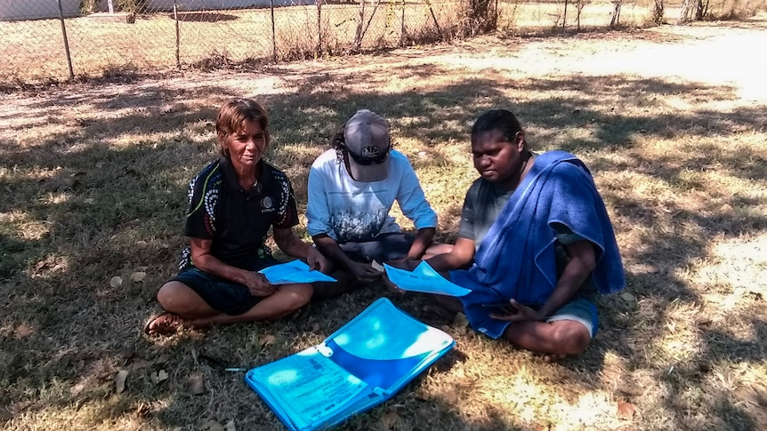 A woman sits on the ground with two other people and reading material. 