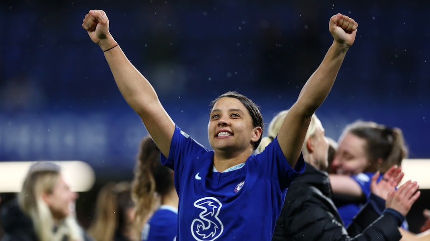 A soccer player wearing blue holds her arms in the air and smiles after winning a game