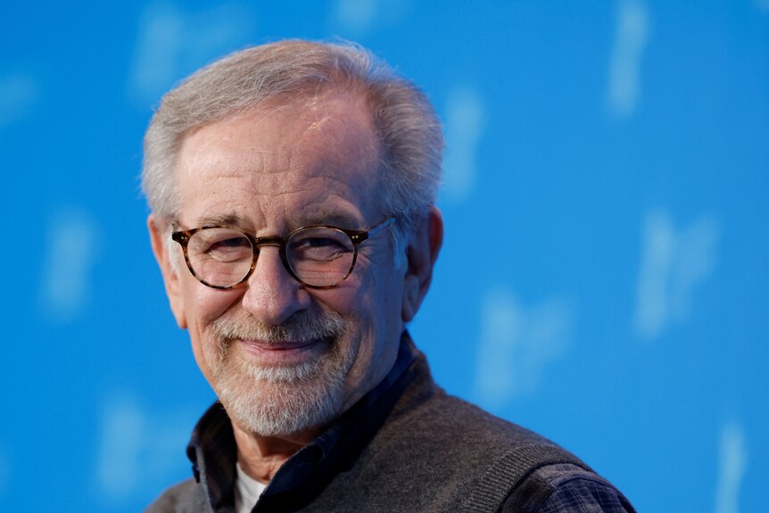 A close up of Steven Spielberg smiling in front of a blue background.