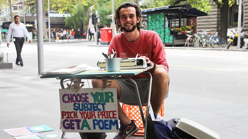 A young man sits behind a table with a typewriter and a sign reading choose your subject pick your price get a poem.