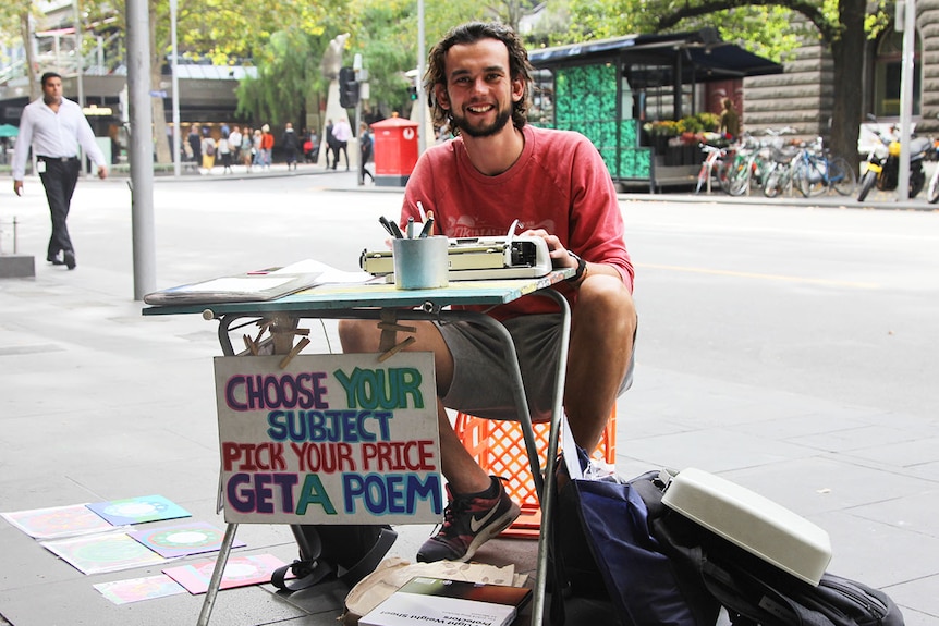 A young man sits behind a table with a typewriter and a sign reading choose your subject pick your price get a poem.