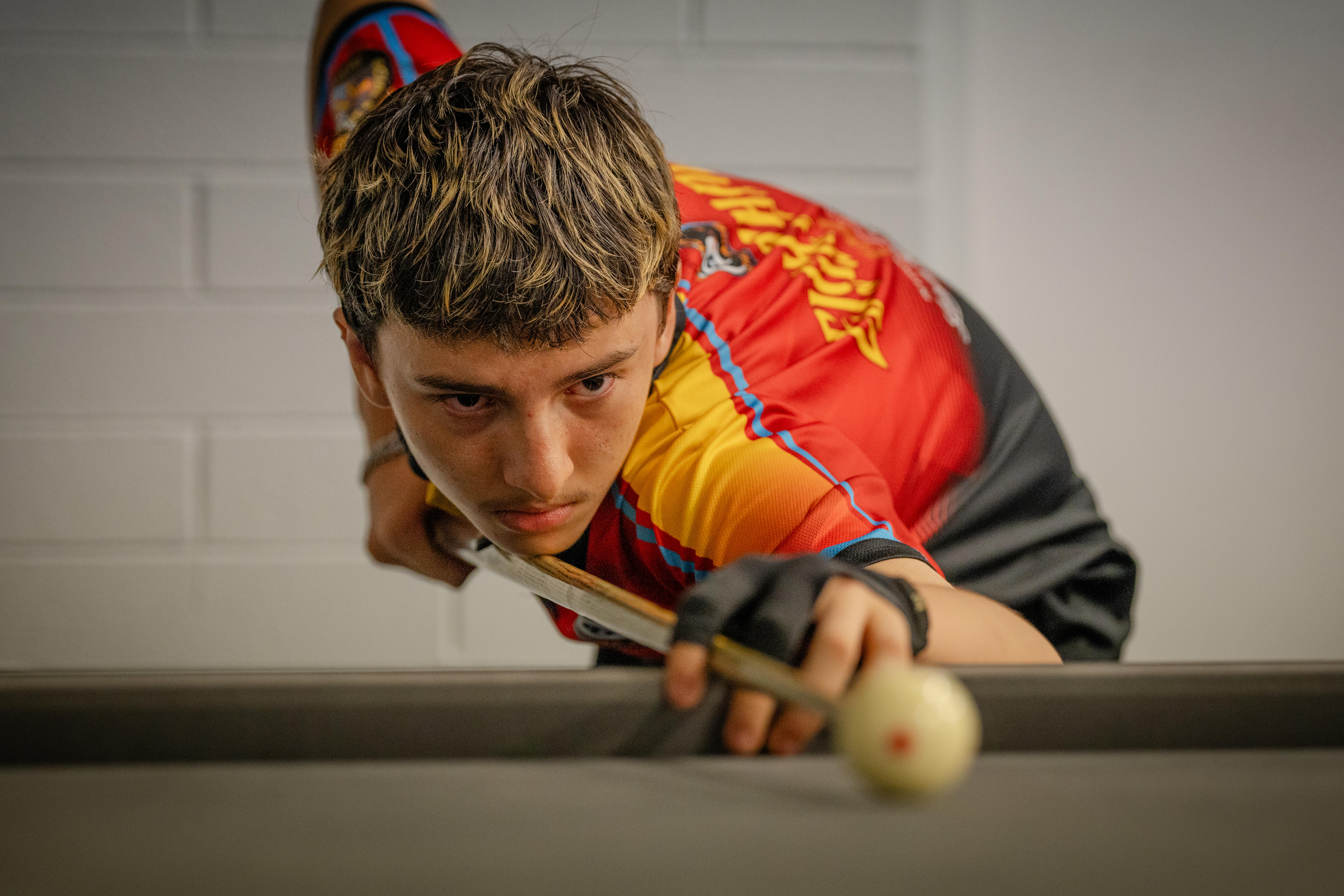 A close-up of Adam Grace leaning over a pool table, lining up his cue with the white ball, a serious expression on his face.
