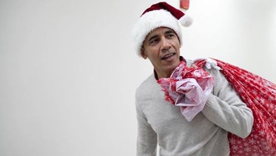 Barrack Obama wearing a Santa hat and carrying a red sack full of presents.