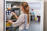 A young woman with long hair looks inside a full fridge containing soft drink cans, sauces, eggs, a saucepan and leftover salad.