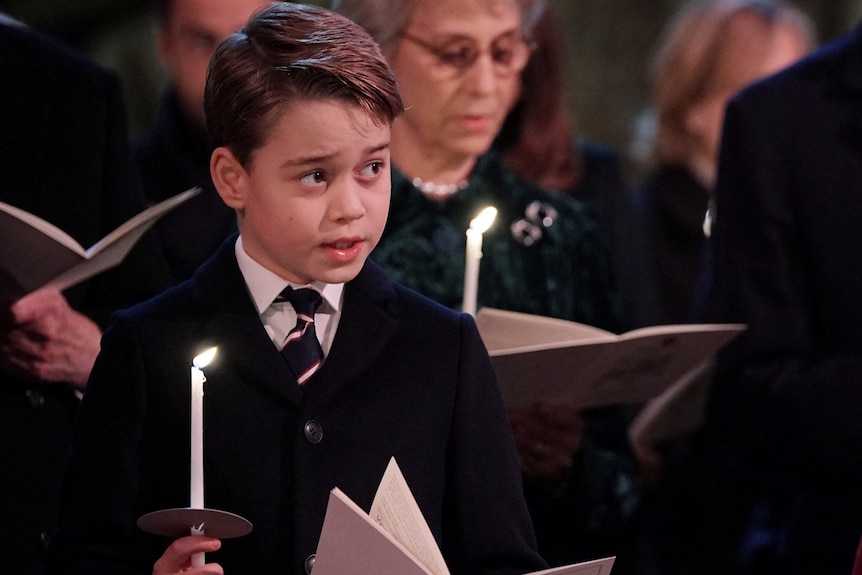 Prince George has his mouth open in song as he holds a candle during a church service. Charlotte looks down at the program 