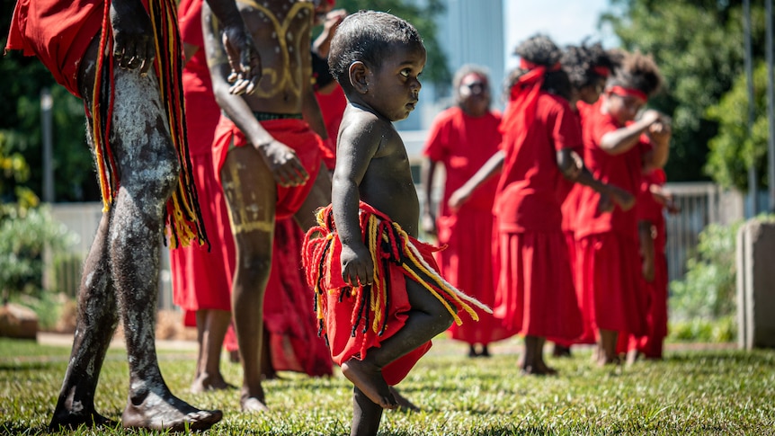 A toddler in traditional Indigenous garb, dancing among some adults.