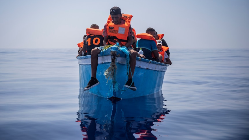 A man sits on the prow of a small boat in a glassy sea.