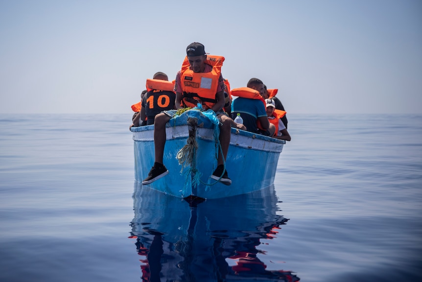 A man sits on the prow of a small boat in a glassy sea.