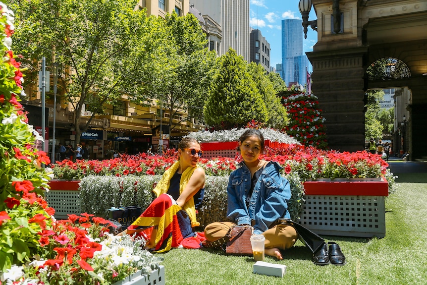 Two women sit on grass in front of red and white garden.