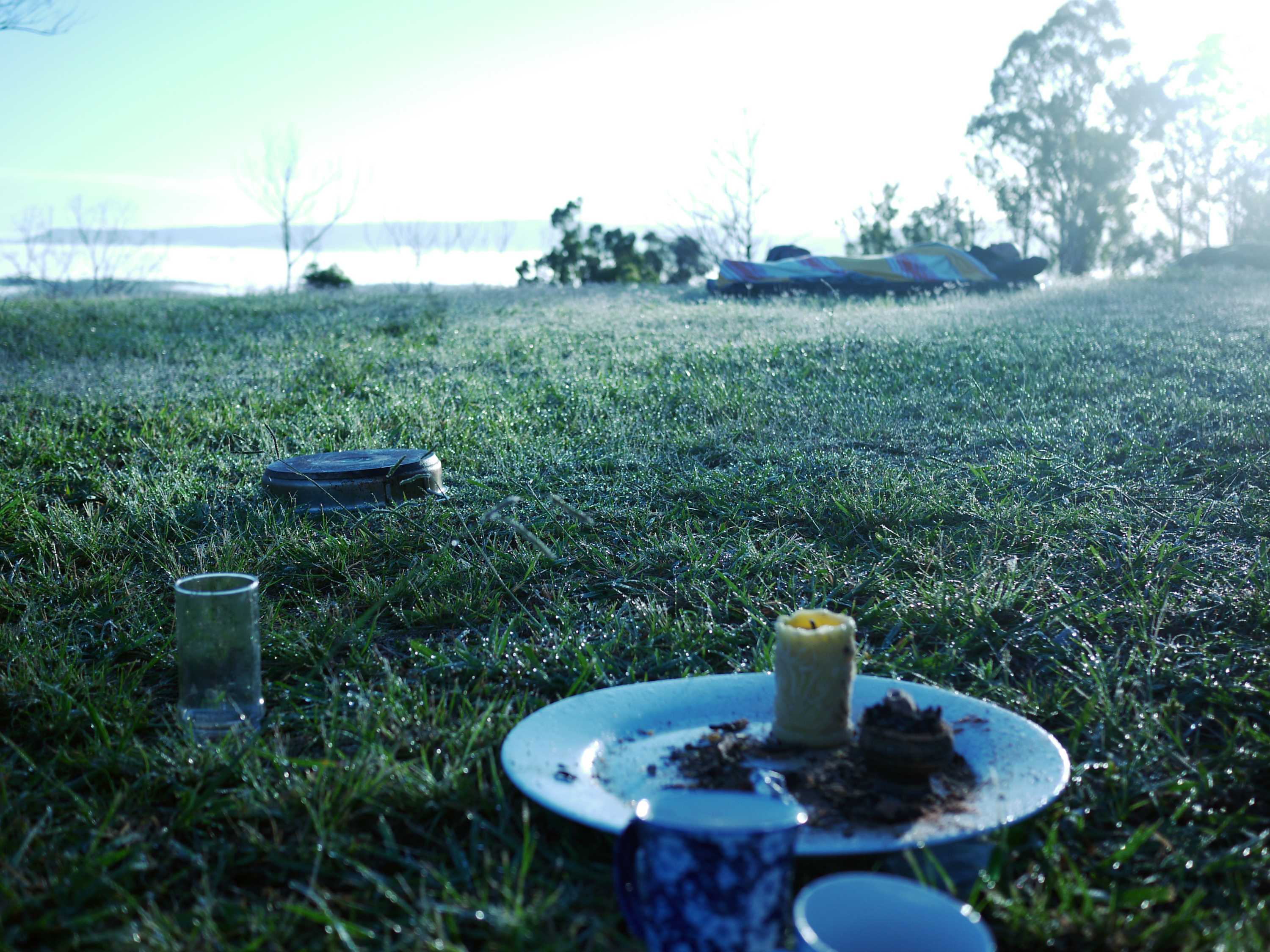 A candle, ashtray, glasses and other items sit on grass, in the background al man reclines under a sheet.