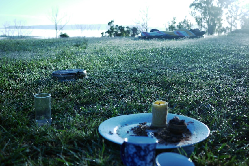 A candle, ashtray, glasses and other items sit on grass, in the background al man reclines under a sheet.