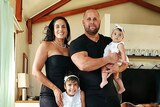Man in black t-shirt holds baby, woman in black dress at his right side, young girl smiles at front of photo.