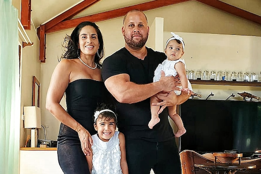 Man in black t-shirt holds baby, woman in black dress at his right side, young girl smiles at front of photo.