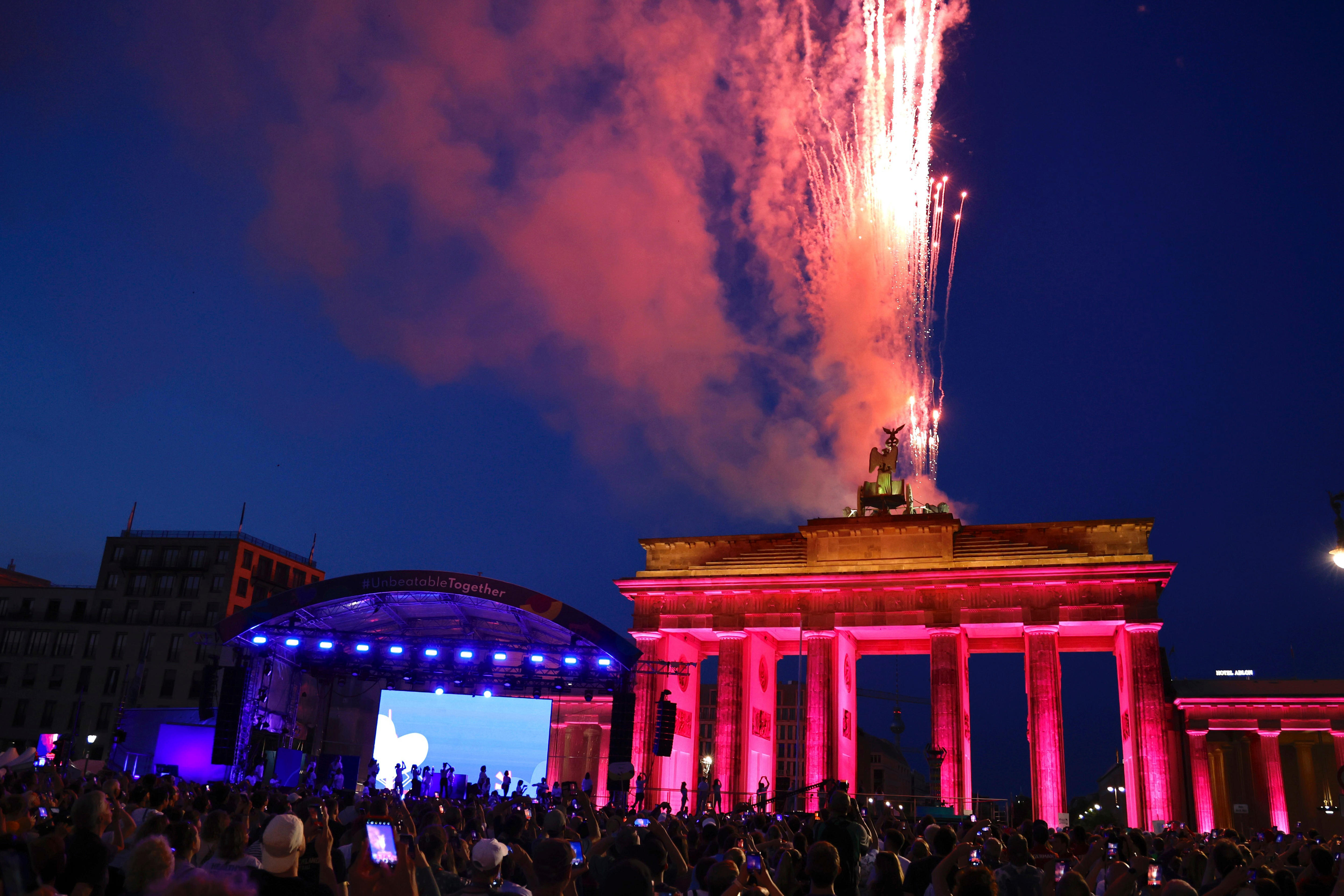 Fireworks burst in the sky over a big German monument, as crowds gather with a big screen in the background.