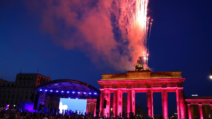 Fireworks burst in the sky over a big German monument, as crowds gather with a big screen in the background.