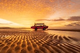 A picture of a bus driving on the beach during an orange sunset