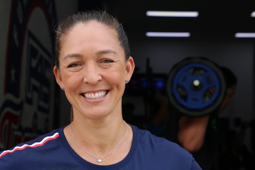 A close-up photo of a woman wearing a t-shirt with someone lifting weights in the background.