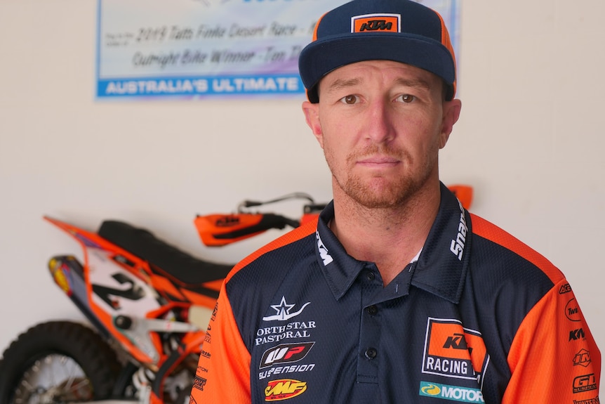 A man wearing his sponsor's hat and t-shirt stands in front of a motorbike looking solemn.