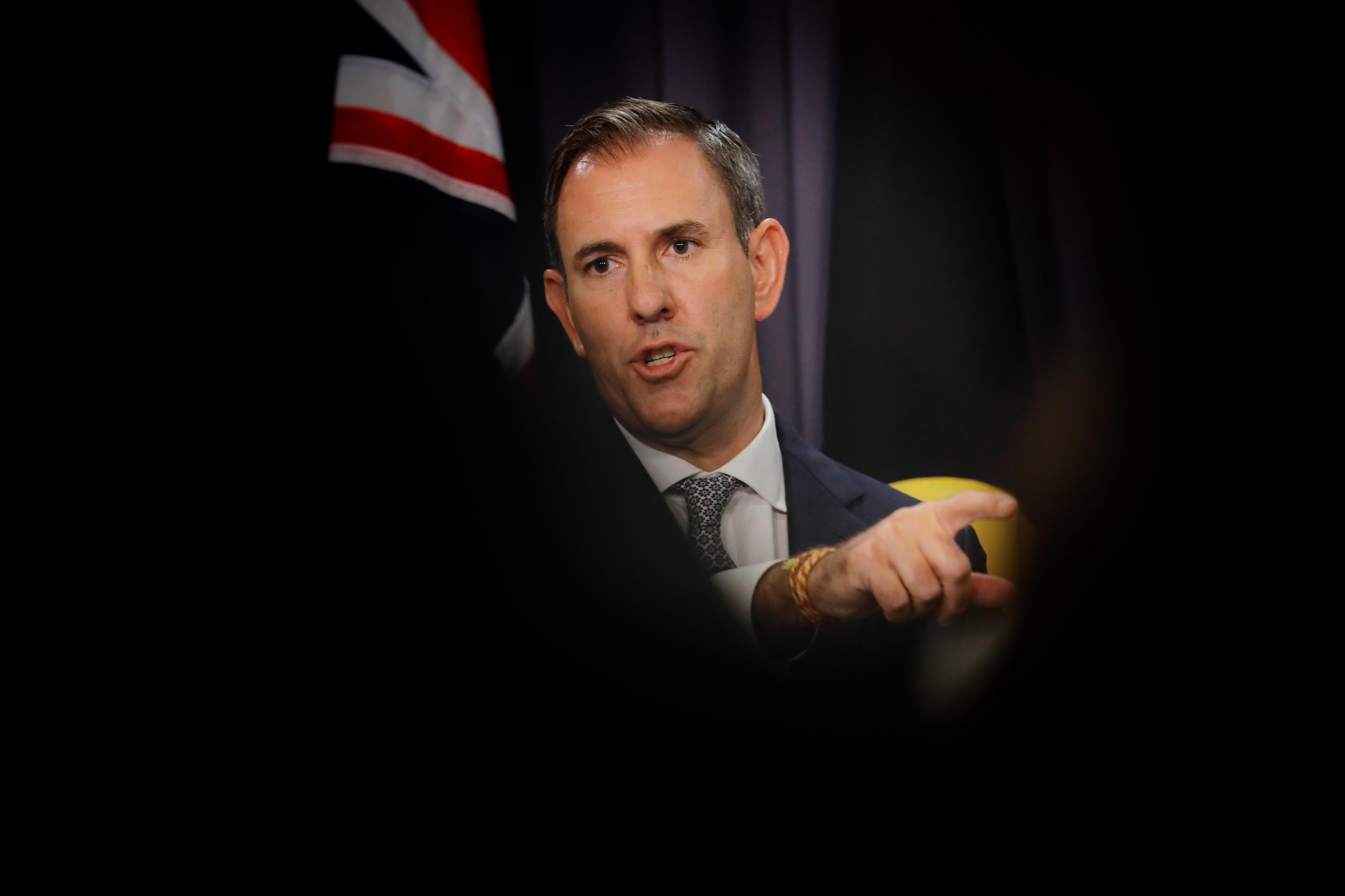 A middle-aged white man in a suit points as he stands in front of an Australian flag, with the photo shot between dark shadows.