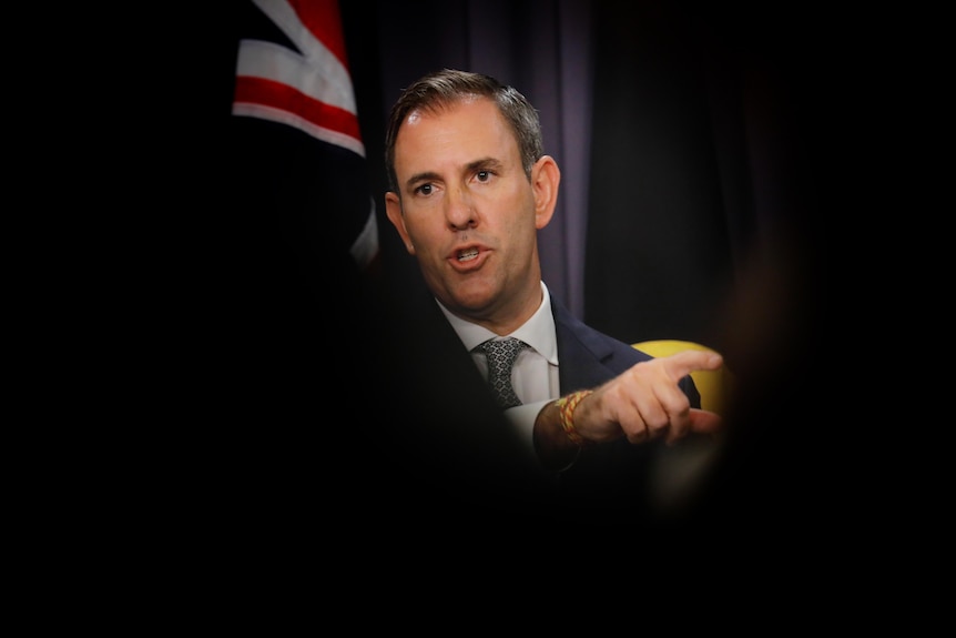 A middle-aged white man in a suit points as he stands in front of an Australian flag, with the photo shot between dark shadows.