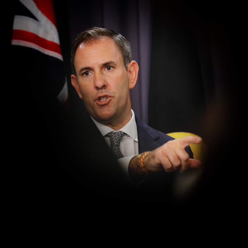 A middle-aged white man in a suit points as he stands in front of an Australian flag, with the photo shot between dark shadows.