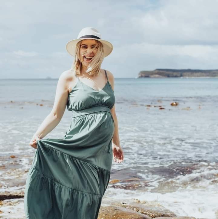 A pregnant woman wearing a green dress and hat stands on rocks on the edge of a beach