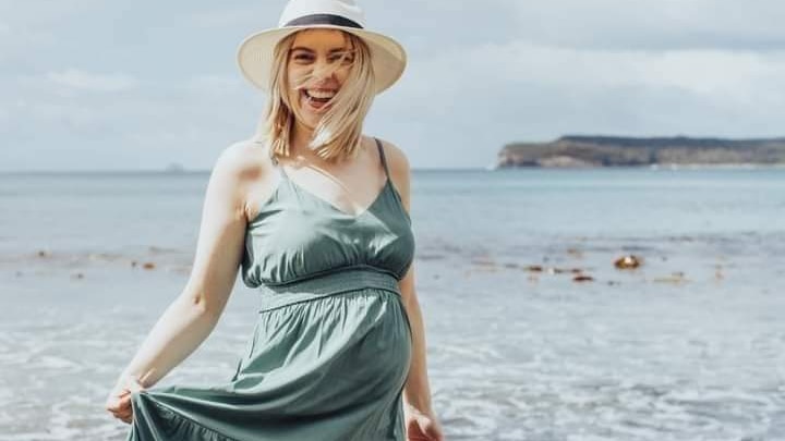 A pregnant woman wearing a green dress and hat stands on rocks on the edge of a beach