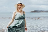 A pregnant woman wearing a green dress and hat stands on rocks on the edge of a beach