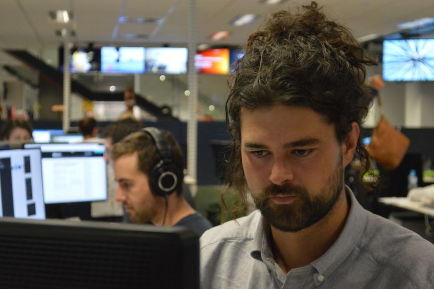 Mantesso sitting at desk in newsroom with man in background wearing headphones.