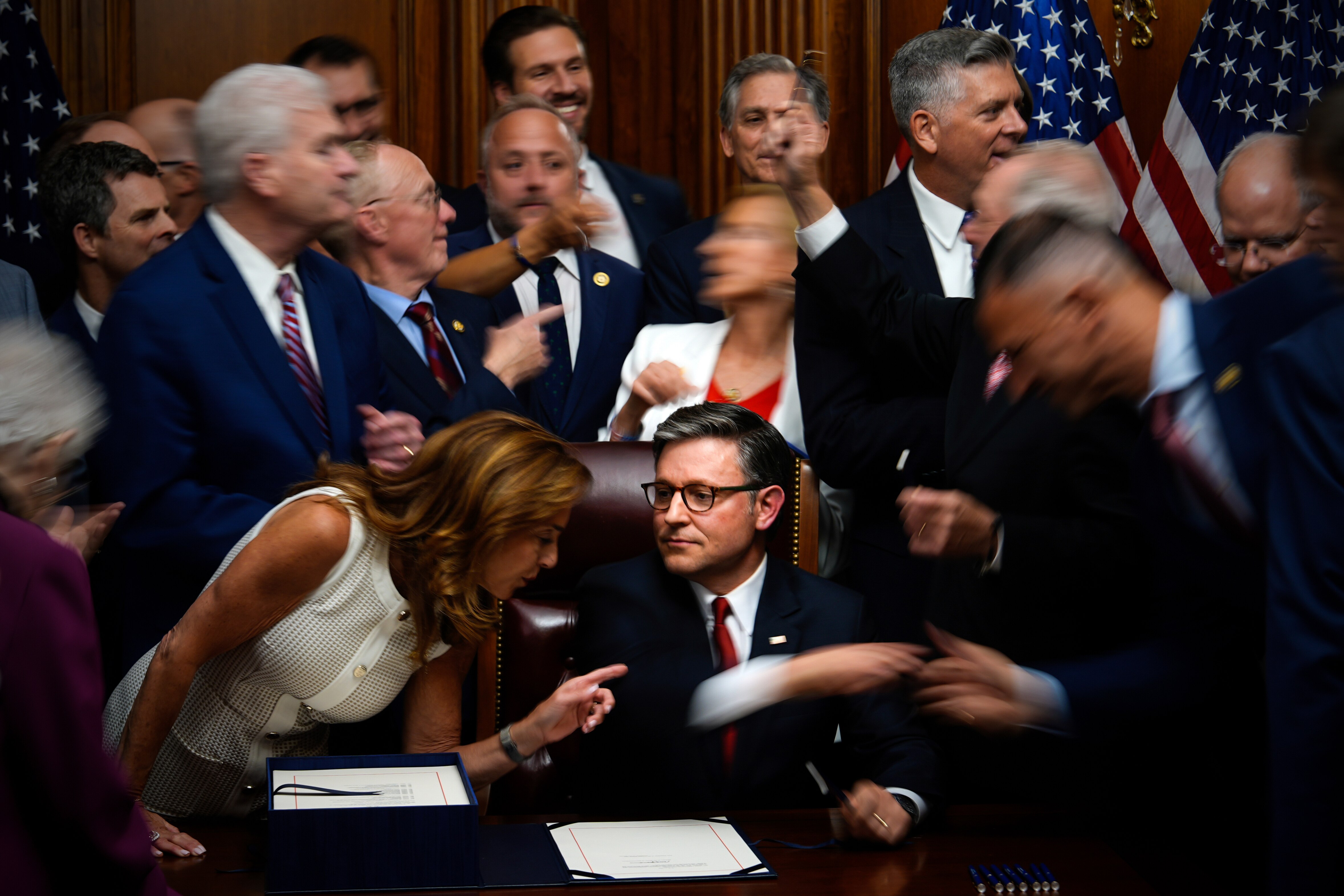 A crowd of people stand around a man in a suit sitting at a desk