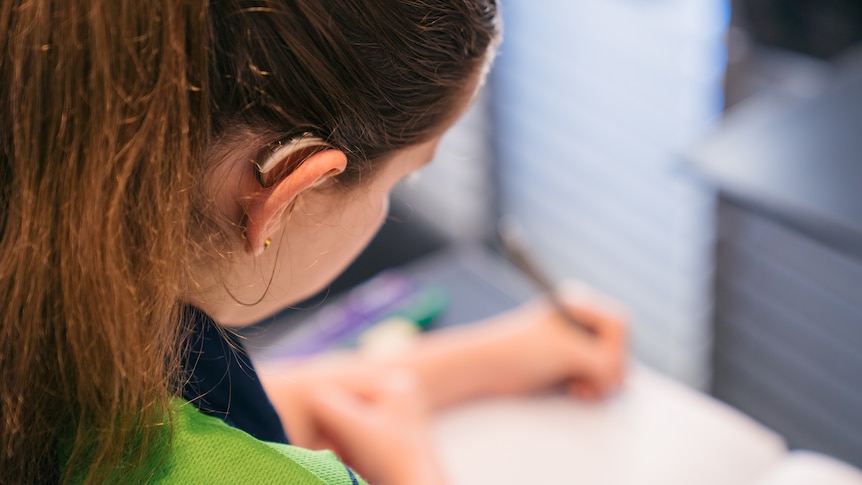 A close-up shot over the shoulder of a young girl sitting at a table writing in a notepad.