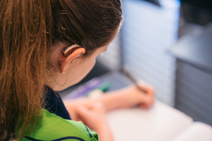 A close-up shot over the shoulder of a young girl sitting at a table writing in a notepad.