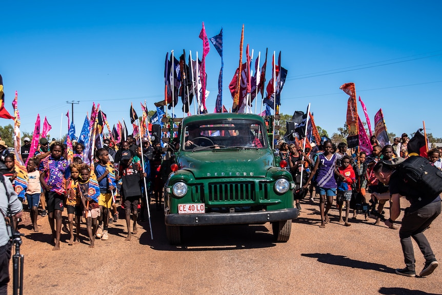 A green truck is flanked by children holding flags and marching. 