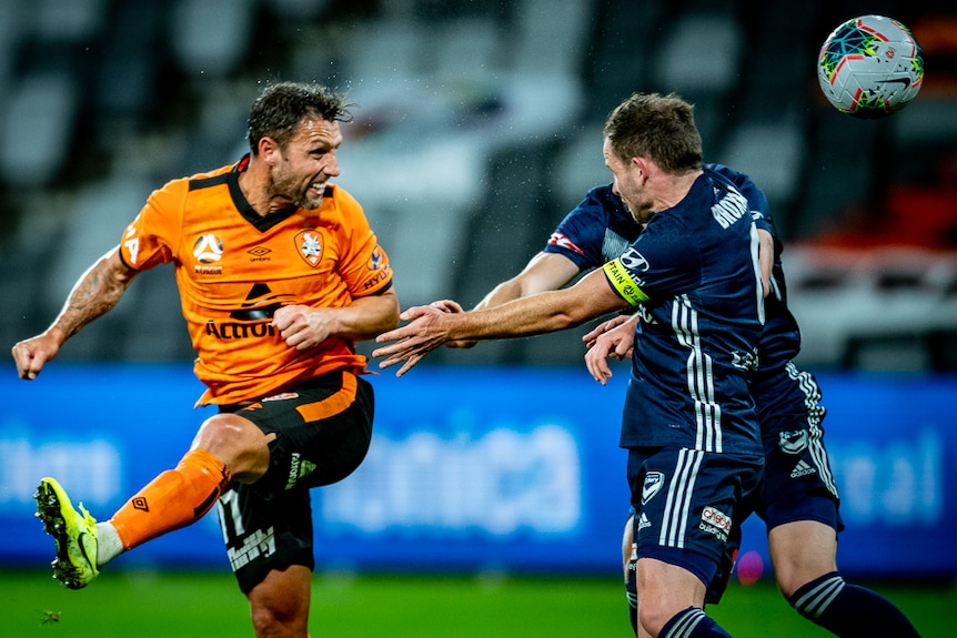 Scott McDonald clenches his teeth and watches the ball as it goes away from his past a player in a blue shirt