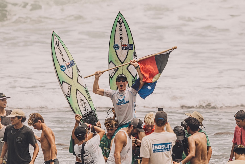 A surfer on people's shoulders is brought up the beach with surfboards held up behind him