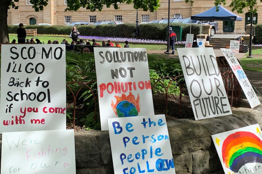 Seven placards are seen resting on and on top of a wall, with a small crowd of protesters seen behind.