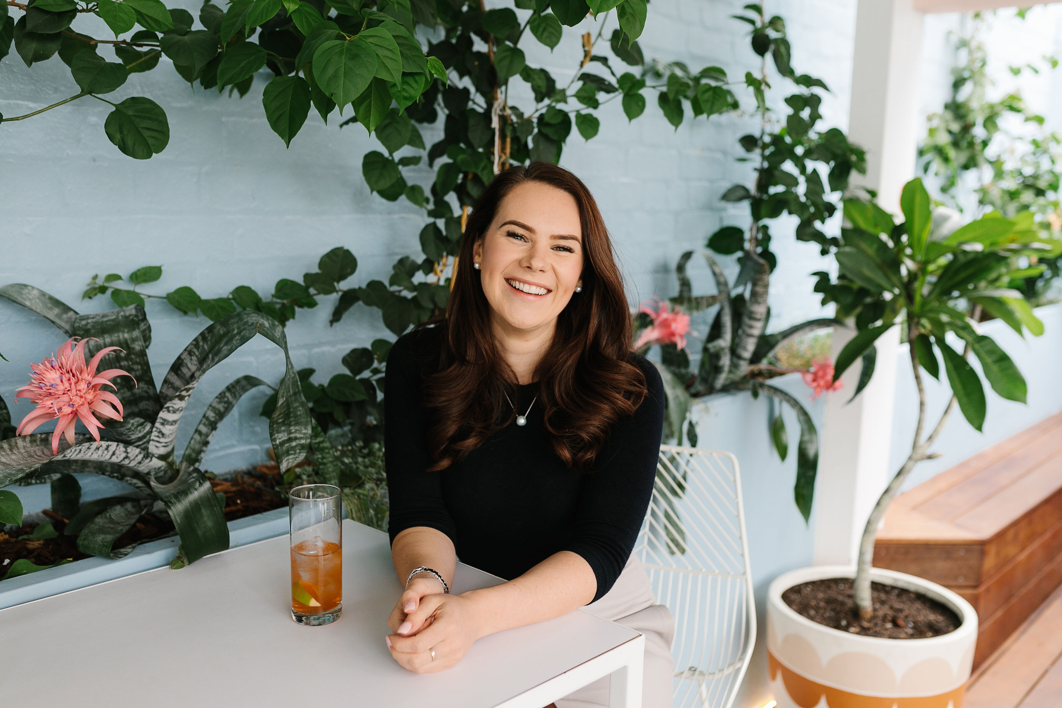 Victoria Devine sits at a table outside with a drink and plants climbing a blue wall behind her. 