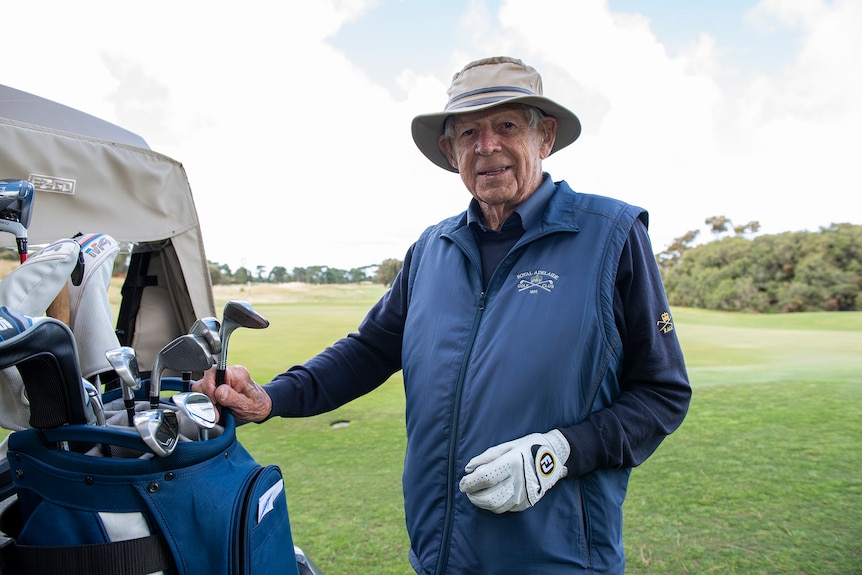 A man in a hat stands next to a golf buggy with golf clubs