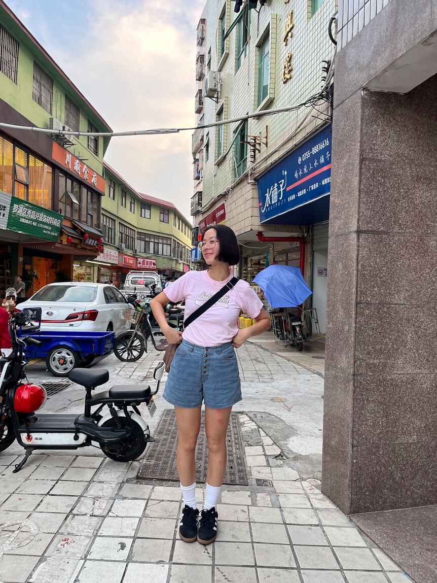 Writer Maggie Zhou on a street in Shenzhen, China in a pink t-shirt, denim shorts, crossbody bag and black sneakers.