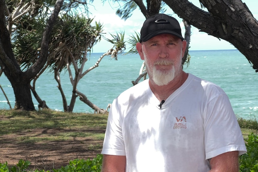 A man with a white beard and wearing a white shirt and black cap standing in front of the ocean